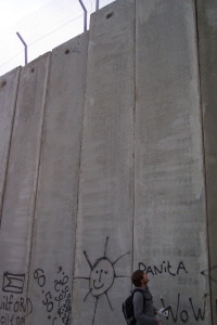 Die Mauer in Bethlehem, Palästina, die lachende, gemalte Sonne wirkt groß neben einem Menschen, doch die Mauer bleibt riesig und trist.