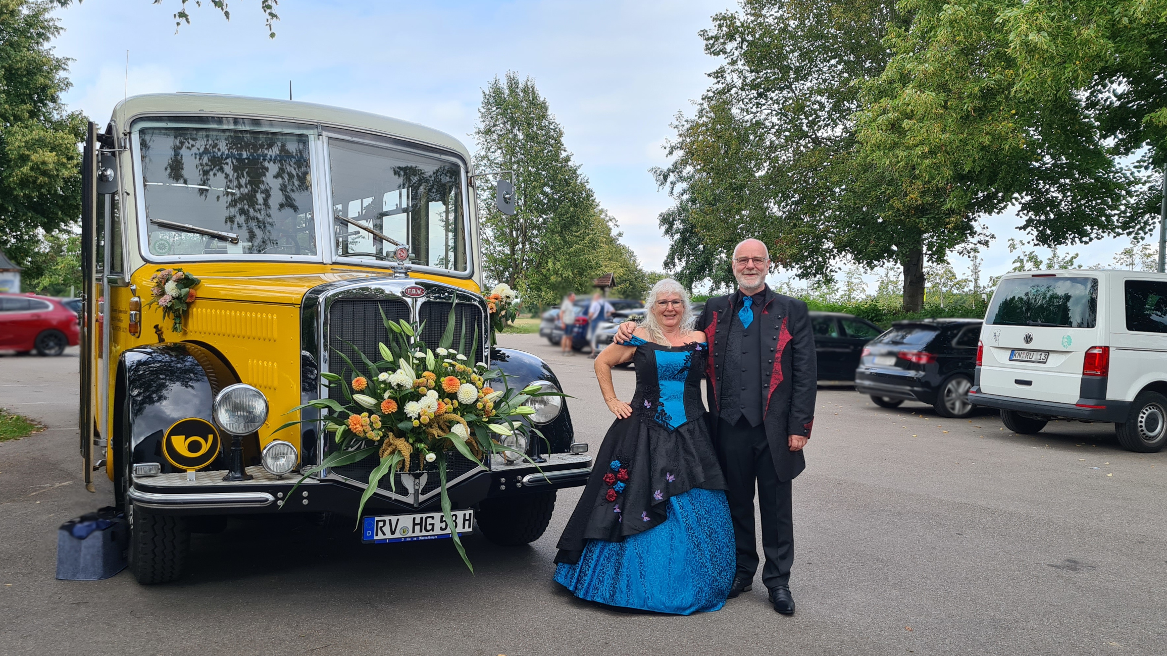 gelber Oldtimer-Postbus mit Blumenschmuck davor ein Paar in Gothic-Outfit in schwarz-petrol