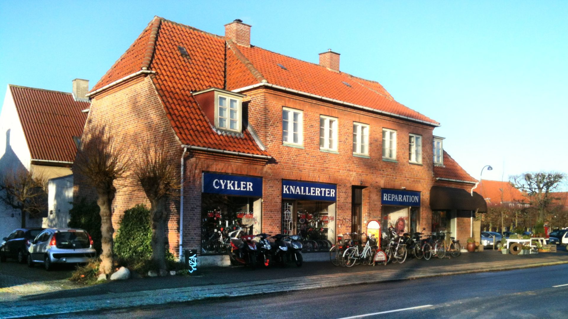 Sonnig-blauer Himmel, ein hübsches rotes Backsteingebäude mit drei Schaufenstern Cykler, Knallerter, Reparation, innen und außen alles voller Zweiräder mit und ohne Motor.